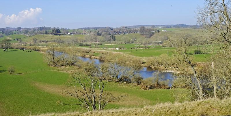 Countryside landscape with a river flowing through green fields and trees, under a clear blue sky.