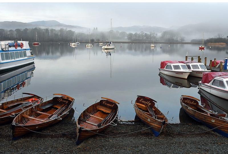 Wooden rowing boats and small motorboats docked on a calm and misty lake, with other various boats floating in the background.