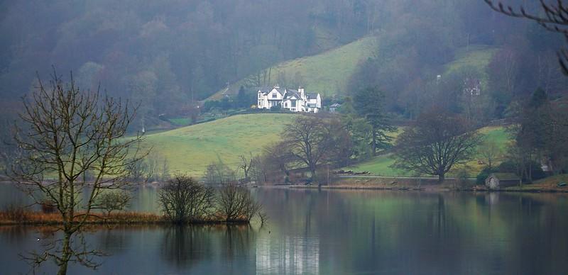 A large white house situated on a lush green hillside overlooking a serene, reflective lake, surrounded by leafless trees.