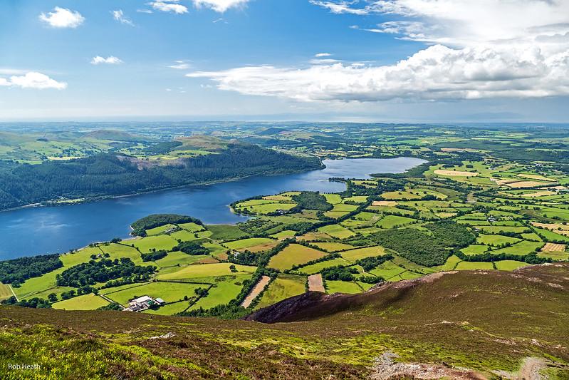 Scenic view of a lake surrounded by lush green fields, forests, and hills under a partly cloudy sky.