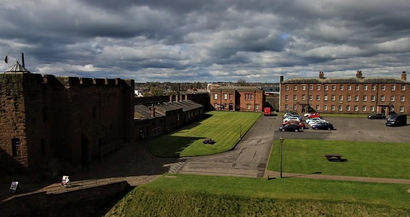 View of Carlisle Castle courtyard, featuring historic stone and brick buildings, green lawns, parked cars, and dramatic, cloudy skies.