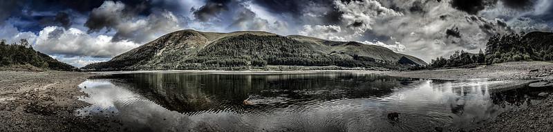 Panoramic view of a calm lake surrounded by rocky shores and forested hills, with a dramatic cloudy sky overhead.