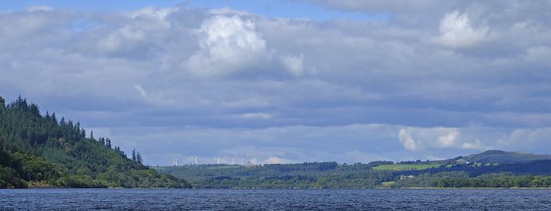 Lake surrounded by forested hills under a cloudy sky with wind turbines in the distance.