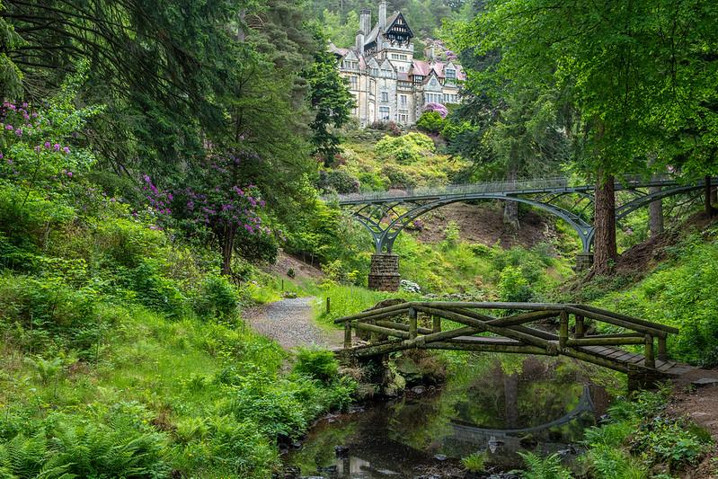 A path and wooden bridge over a stream in a lush green forest with a large manor house and a larger arched stone bridge in the background.