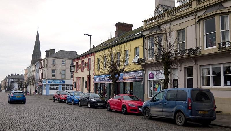 Street view of a row of shops and cars parked alongside an old cobblestone road with a church spire in the background.