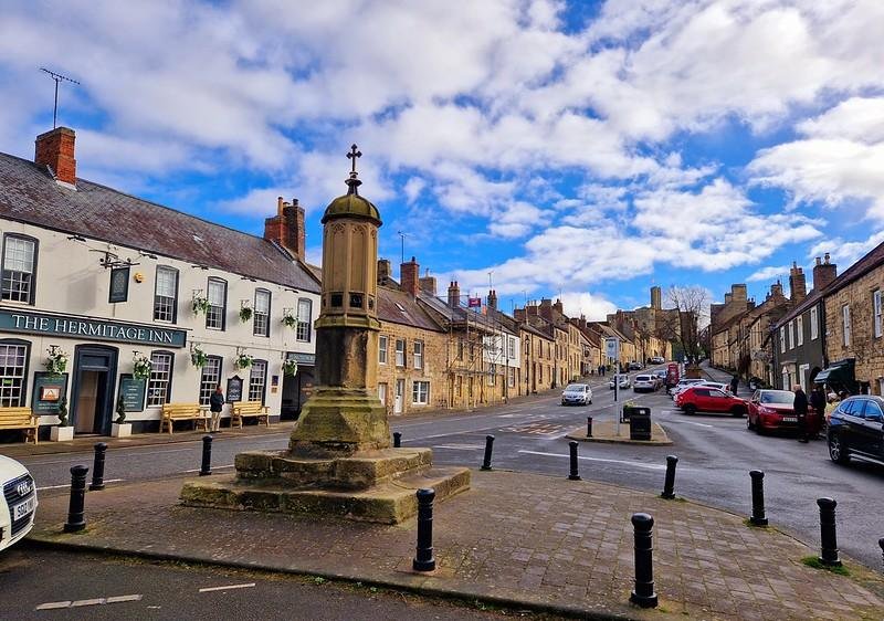 Town square with a stone memorial in the centre, flanked by the Hermitage Inn on the left and a variety of stone-built houses and cars on the right under a partly cloudy sky.