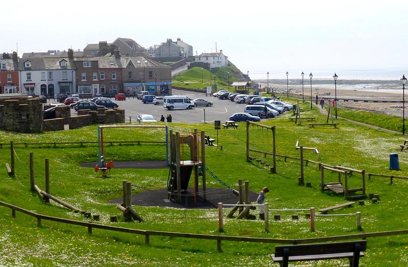 Playground in a grassy park, surrounded by houses, a car park, and a sea in the background.