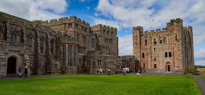 Medieval castle building with a large green lawn in front, featuring a mix of architectural styles including a tall square tower and ornate stone walls, with people walking and enjoying the area under a partly cloudy sky.