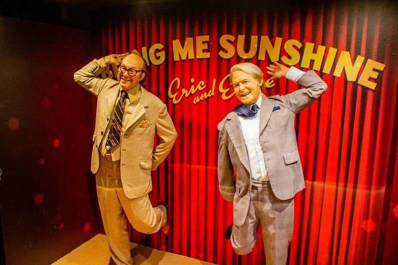 Wax figures of two men wearing suits, posing with exaggerated gestures in front of a red curtain background with the words “Bring Me Sunshine” above them.