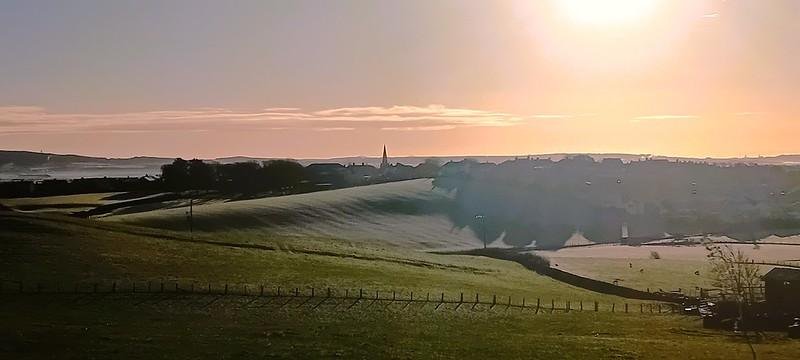 Rolling fields with early morning sunlight, distant town and church spire under a clear sky.