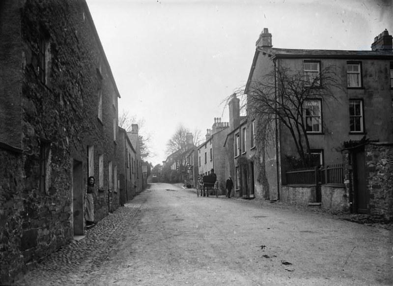 A vintage photograph of a quiet, cobblestone street lined with stone buildings on both sides, with a few people and a horse-drawn carriage in the distance.