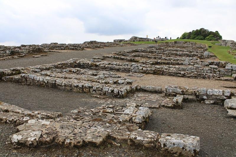 Ancient stone ruins with low walls on a slightly hilly terrain under a cloudy sky.