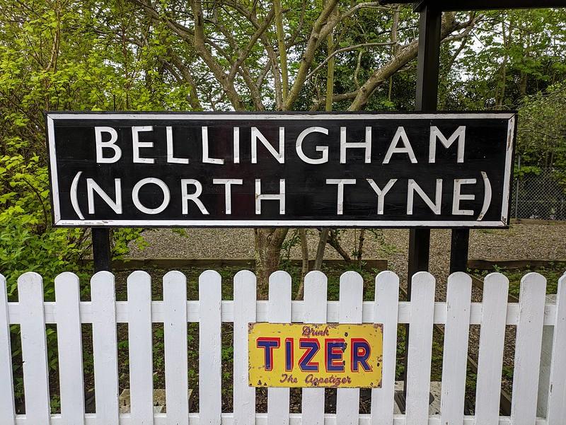 A sign reading "Bellingham (North Tyne)" behind a white picket fence with a vintage Tizer drink advertisement.