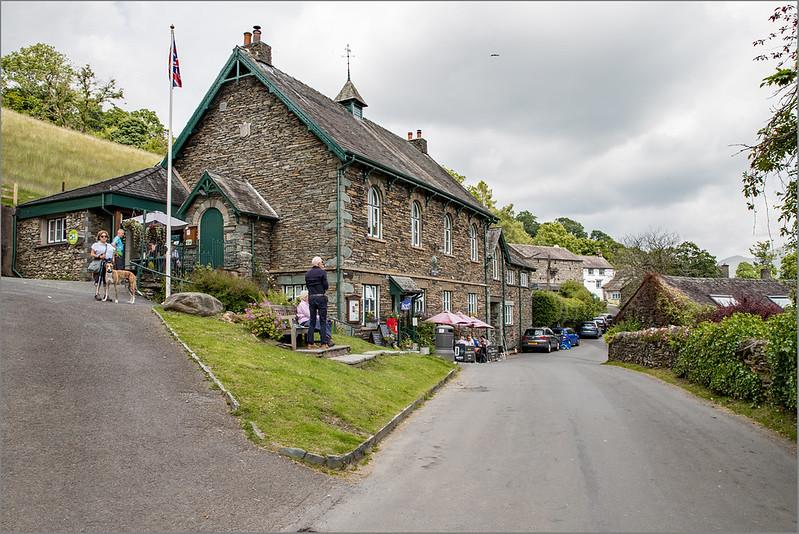 A street with a stone building on a grassy hill, people walking with a dog, a Union Jack flag, and cars parked along the side.