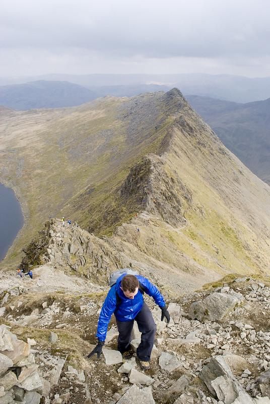 Helvellyn, Cumbria | A hiker in a blue jacket climbs a rocky mountain ridge with a steep drop on one side, under a cloudy sky.