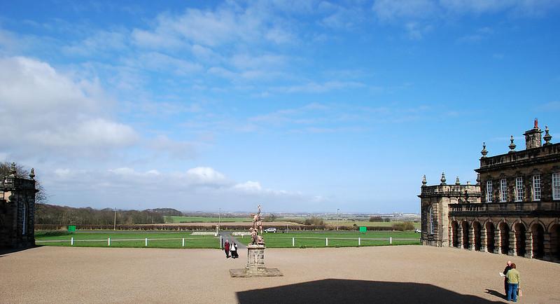 Seaton Delaval | Large open courtyard with a statue in the centre, flanked by historic buildings on either side, under a blue sky with scattered clouds.