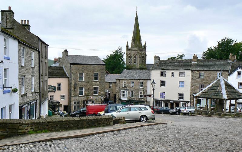 Alston Town square with cobblestone streets, parked cars, historic buildings, and a church with a tall steeple in the background.