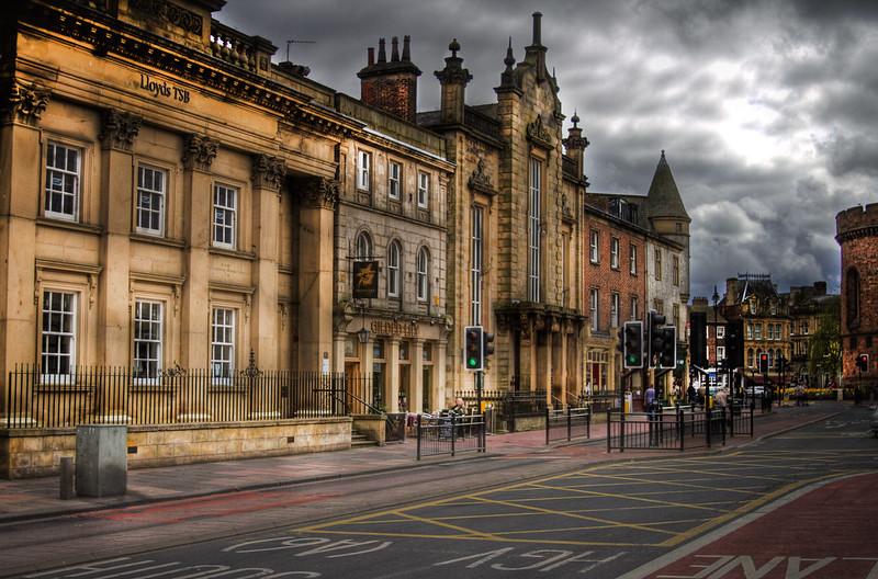 Historic buildings along a street in York, England, featuring ornate architectural details and traffic lights, under a cloudy sky.