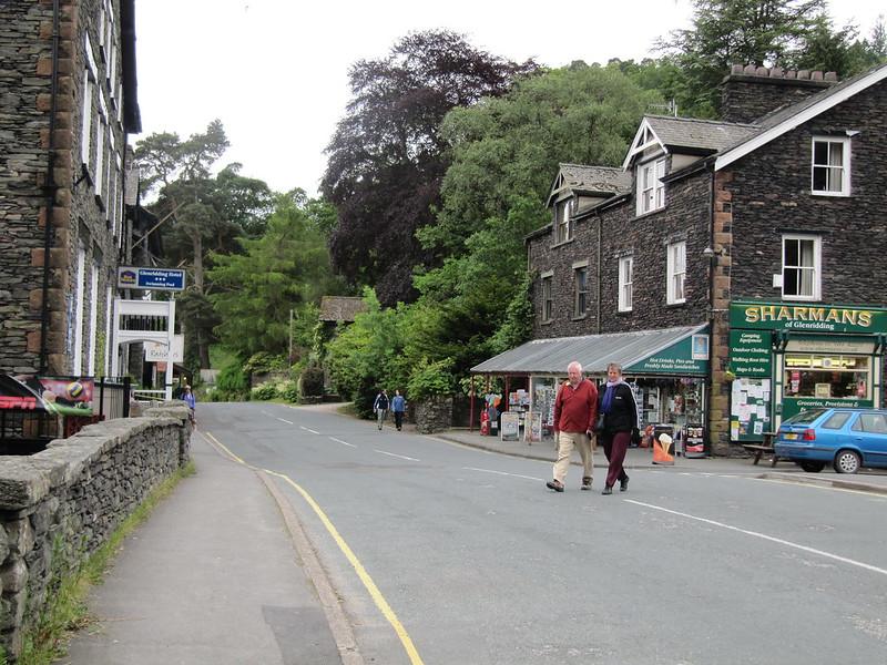 A street scene in a village with stone buildings, a shop called "SHARMANS," people walking on the pavement, and cars parked alongside the road. Trees and greenery are in the background.