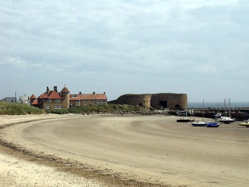 Beadnell - Northumberland | Sandy beach with scattered boats, a row of houses, and a large stone fortification in the background.