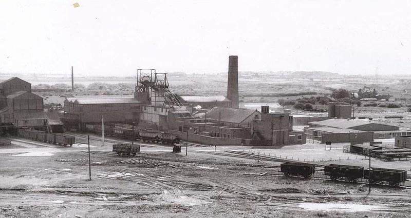 Choppington Colliery, Northumberland (1963) | Black and white photograph of a large industrial coal mine, featuring a headframe, chimneys, several buildings, and rail carts on tracks.