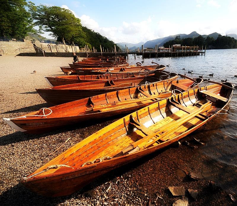 Wooden rowboats lined up on a pebbly shore beside a lake, with a dock and a mountainous landscape in the background.