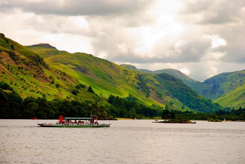 A boat sailing on a lake surrounded by lush green hills and partly cloudy skies.