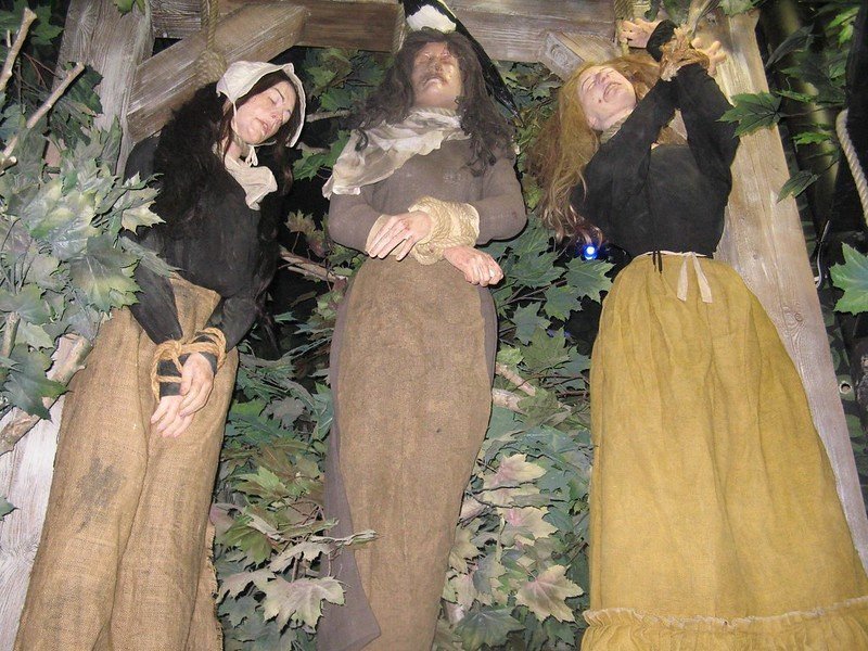 Three mannequins dressed in historical clothing, posed as if hanging from a wooden gallows amidst leafy branches.