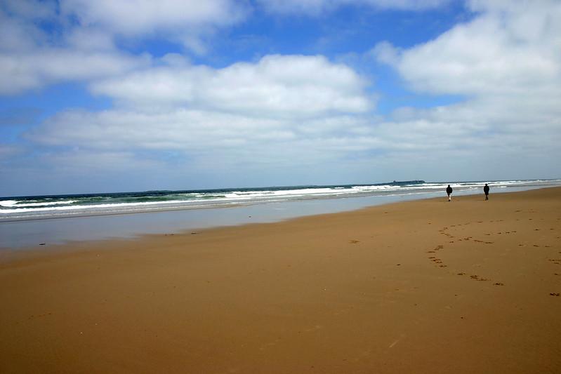 Bamburgh beach | A vast sandy beach with two people walking along the shore, under a partly cloudy blue sky.