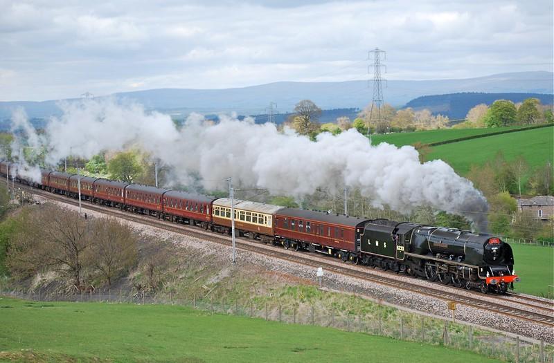 A steam locomotive pulling a long line of passenger carriages, emitting clouds of white smoke, travels through a countryside landscape with green fields and rolling hills in the background.