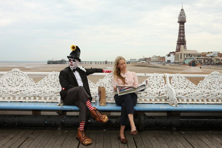 A person dressed as a clown and a woman reading a newspaper sit on a bench by the seaside, with a beach and a tower visible in the background.