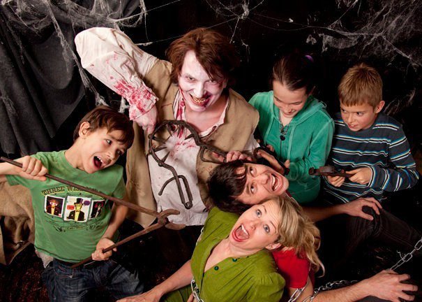 A group of people with frightened expressions, surrounded by fake cobwebs and a person in zombie makeup, posing in a Halloween-themed setting.