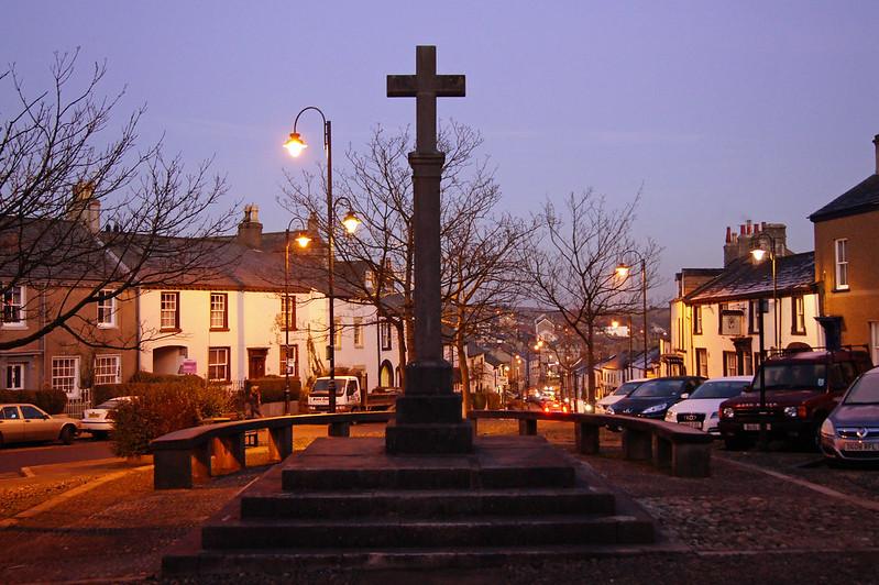 A stone cross monument in the centre of a village square, surrounded by residential houses and streetlights at dusk.