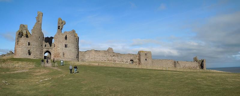 Ruins of Dunstanburgh Castle on a grassy hill under a blue sky.