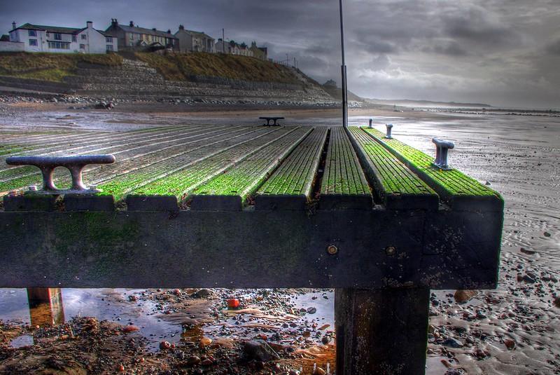 Old wooden pier with green algae on the surface, small houses on a hill in the background, cloudy sky, and sandy beach with scattered stones.