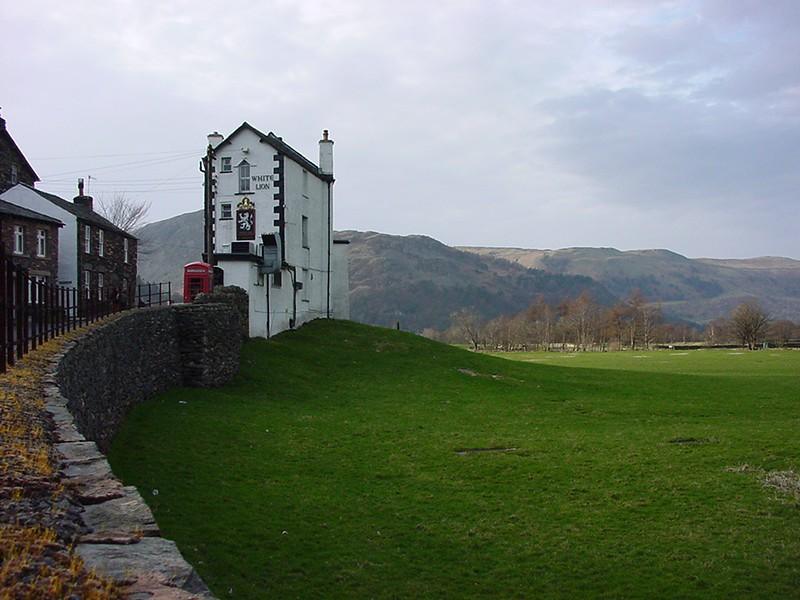 A white building with a sign that reads "The White Lion Inn" on its side, with a red phone box nearby. There is a stone wall to the left, green fields in the foreground, and hills in the background.