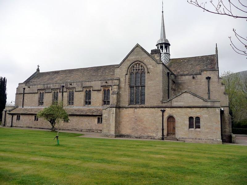 Stone church with arched windows and a small spire, situated on a grass lawn with a tree in the foreground.