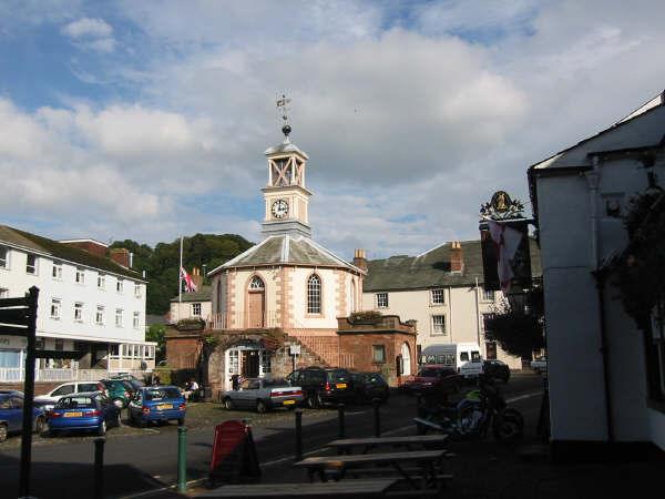 Town square with historic building featuring a clock tower, parked cars, and surrounding structures.