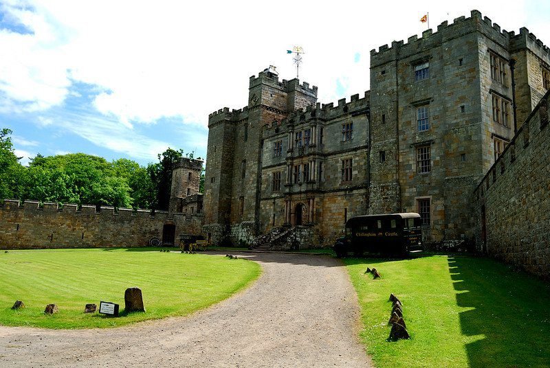 A historic stone castle with a large central building and two towers, surrounded by a grassy courtyard and a stone wall.