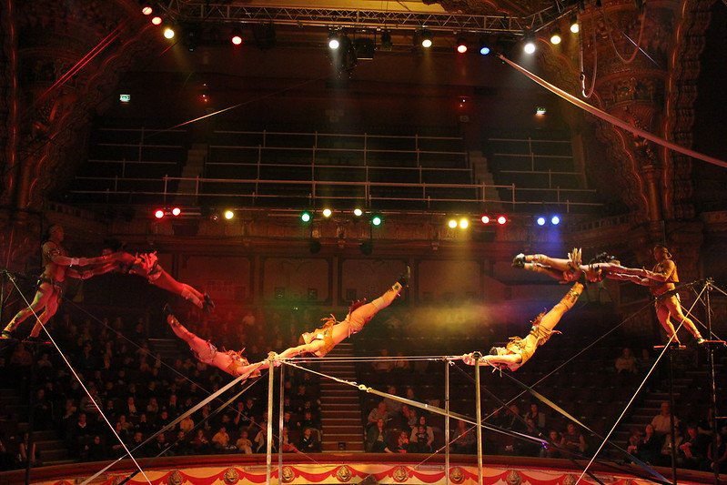 Acrobats performing a daring aerial act on ropes, with an audience watching in a dimly lit arena.