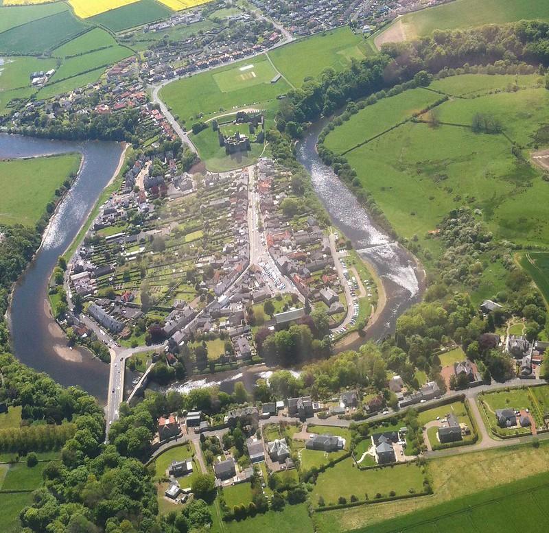 Warkworth, Aerial view of a small town with a river flowing through it, surrounded by green fields and residential areas.