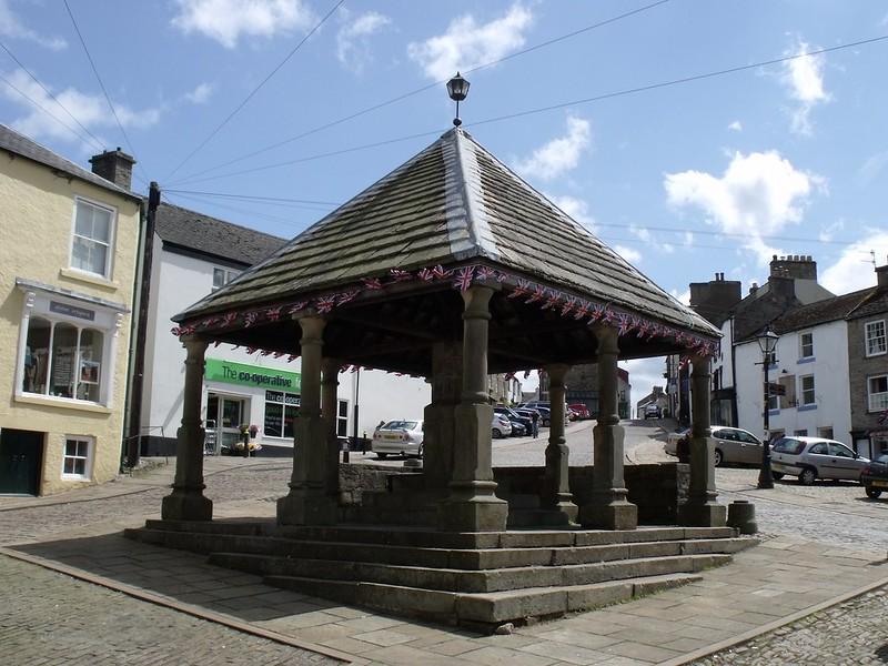 Stone market shelter with a slate roof, Union Jack bunting, and surrounding street view with shops and parked cars.