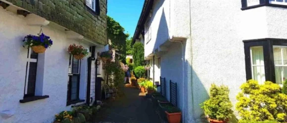 Narrow alleyway between charming white cottages with hanging flower baskets and potted plants, under a clear blue sky.