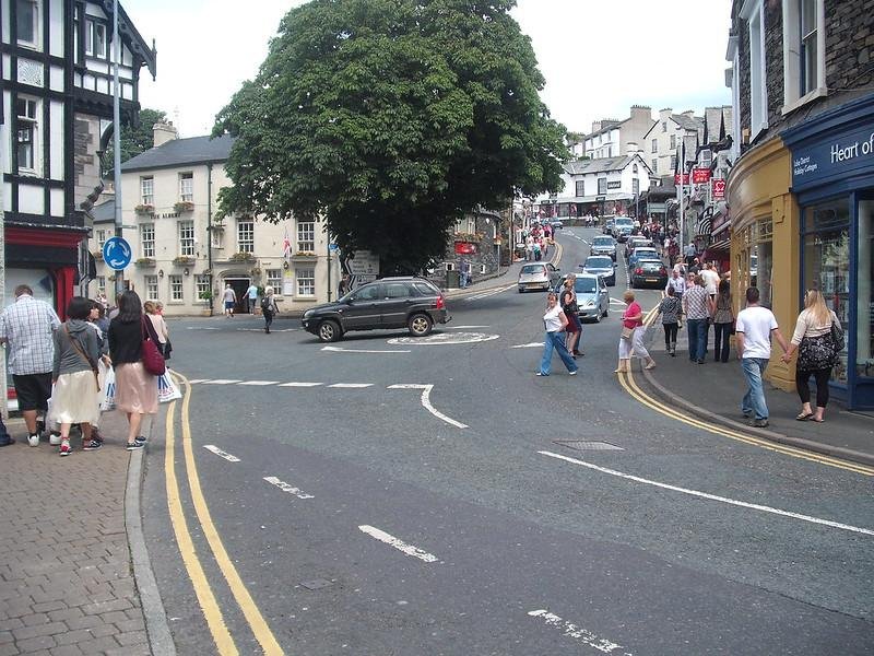 Windermere Town | A street scene in a small town with a tree in the middle of a roundabout, people walking on sidewalks, and cars on the road. Shops and buildings line the street as it slopes upward.