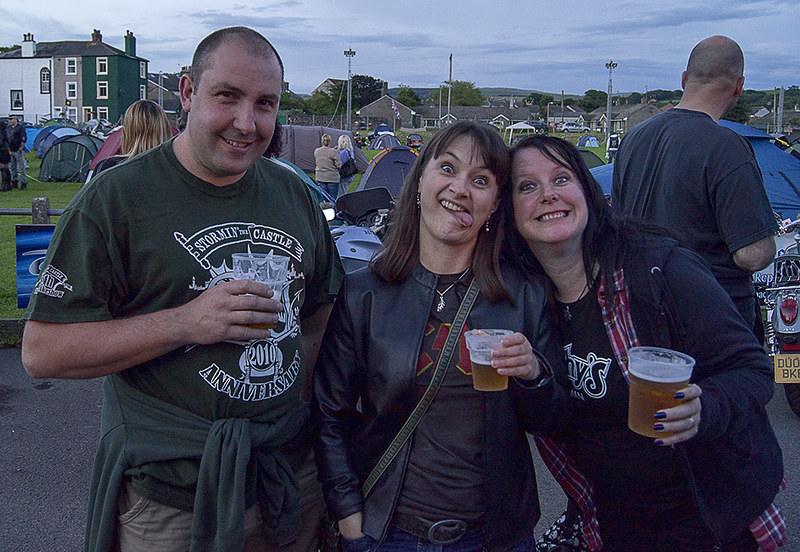 Three people at an outdoor event holding beer, one of them making a silly face. Tents and additional people are visible in the background.