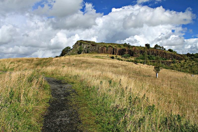 A winding path through tall grass leads towards a rocky hill under a partly cloudy sky.