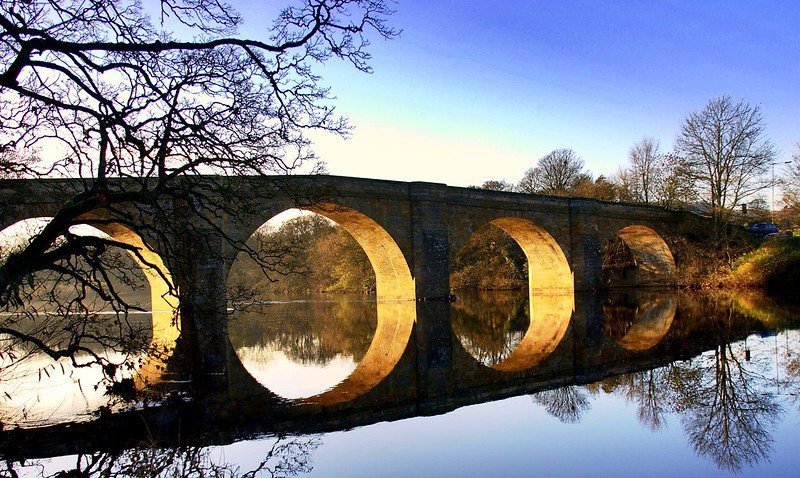 Stone bridge with archways reflecting on a calm river, surrounded by bare trees under a clear blue sky.