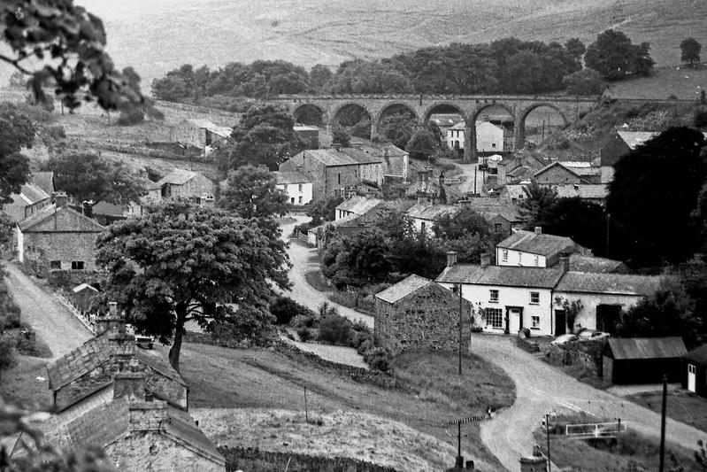 Crosby Garrett Cumbria 1981 | Black and white photograph of a quaint village nestled in the countryside, featuring stone buildings, winding roads, and an arched railway viaduct in the background.