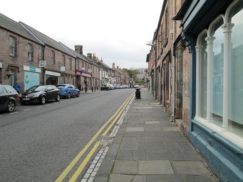 Empty street lined with stone buildings, parked cars on the left, and a paved sidewalk on the right.