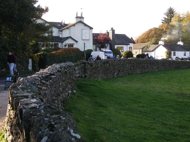 Near Sawrey | Traditional stone cottages and buildings lined along a curved stone wall with lush green grass in the foreground and trees and bushes surrounding.